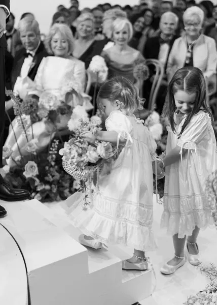 Flower girl moment as two flower girls at ceremony step forward in lace dresses holding baskets, with wedding guests watching behind
