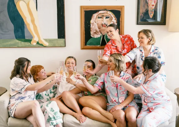 Bridesmaids toast with champagne flutes, wearing printed pajamas on a sofa in a bridal suite with framed artwork and a lamp