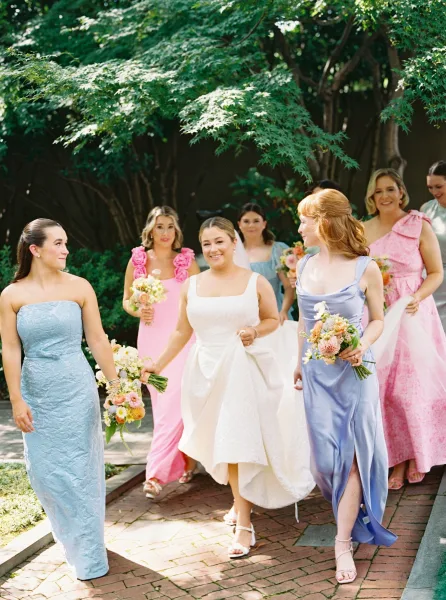 Bride with bridesmaids walking with bride down a sunlit brick garden walkway, holding bouquets as her veil trails behind