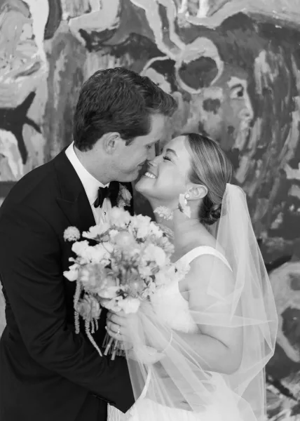 Wedding couple portrait in black and white, bride and groom nose kiss with bouquet and veil against a painted mural wall backdrop