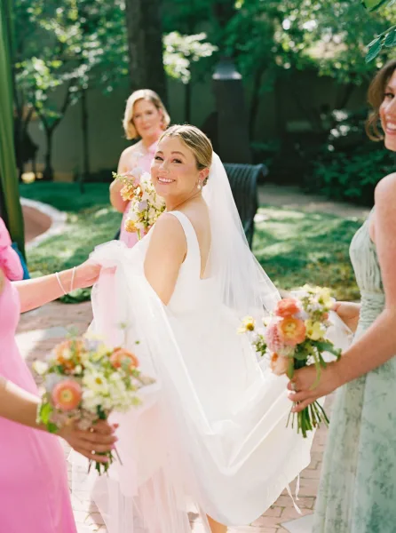 Bride portrait with bride looking back in a long white veil and sleeveless gown, flanked by bridesmaids holding pastel bouquets in a garden courtyard
