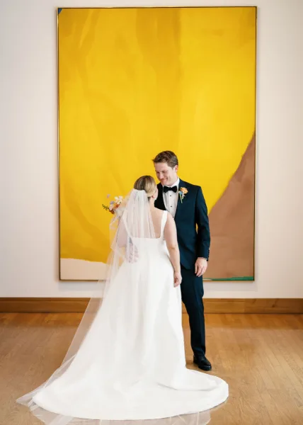 Couple portrait of bride in minimalist dress with cathedral veil and bouquet, groom in tux, beside large abstract painting in gallery