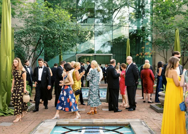 Wedding cocktail hour with wedding guests mingling in tuxedos and formal dresses, holding drinks under string lights in a brick courtyard by a reflecting pool