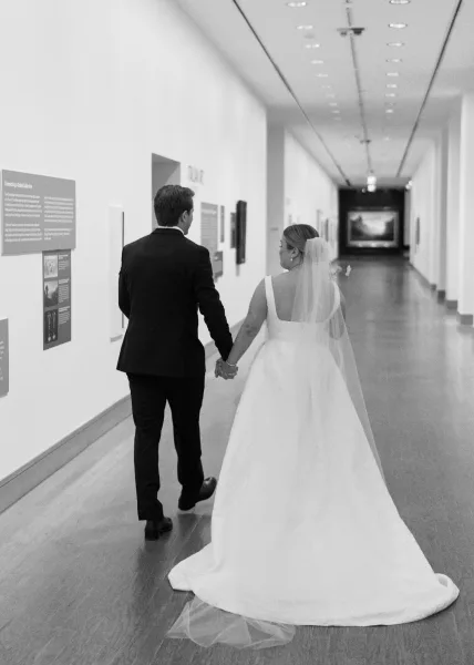 Couple portrait of newlyweds holding hands walking away, bride’s veil and dress train flowing in a framed-art indoor hallway
