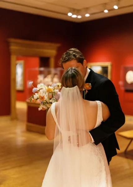 Wedding kiss as bride in flowing veil embraces groom in black suit, holding bouquet in a red-walled art gallery with framed artwork