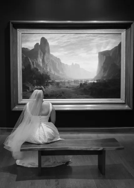 Bridal portrait of a bride sitting on a wooden bench in a black and white bridal portrait, long veil trailing on hardwood before framed art