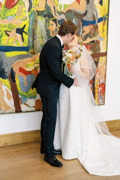 Wedding kiss portrait of bride and groom kissing, bride holding a colorful bouquet with a long veil before abstract wall art indoors