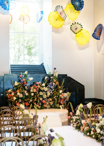 Ceremony altar decor with a wedding ceremony floral installation on a round stage, meadow-style aisle florals and glass wall art behind chairs