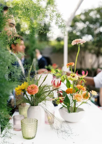 Reception tablescape with wildflower wedding centerpiece in bud and ceramic vases, colored glass votives on a white tablecloth in a garden setting