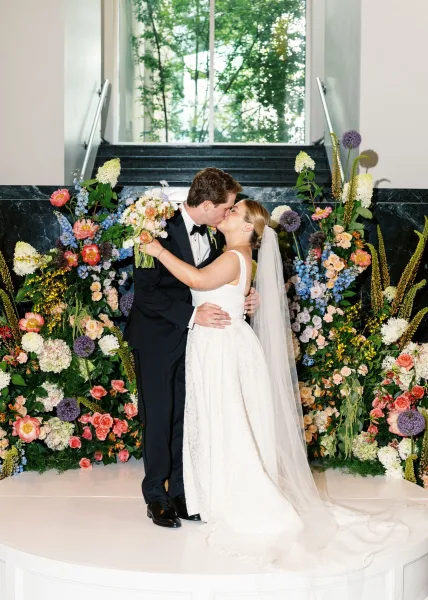 Wedding kiss as bride holds bouquet beside groom in black tuxedo, long veil flowing on an indoor staircase by a large window