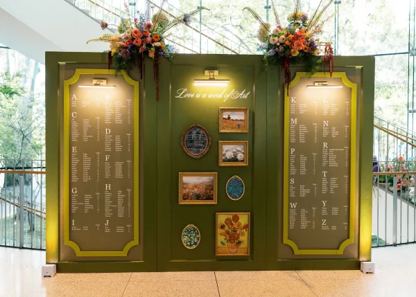 Wedding seating chart with calligraphy lettering on framed panels, topped with florals and picture lights in a glass-walled lobby
