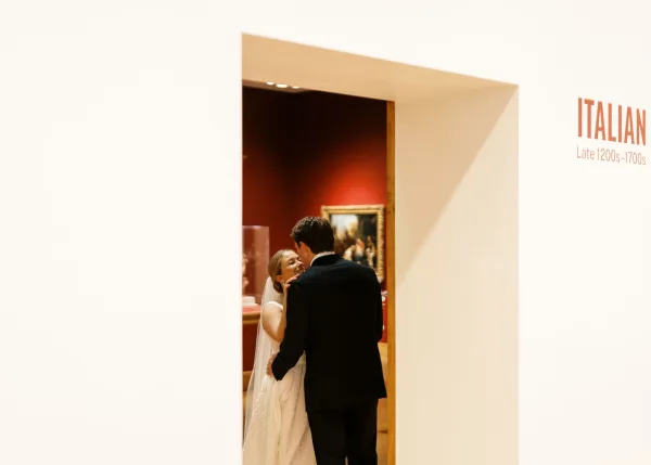 Couple portrait of bride and groom embrace, bride in veil touching his face in a museum gallery with framed artwork on red wall