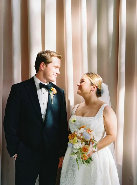 Couple portrait of bride and groom holding hands, bride with veil and bouquet gazing at groom before soft drape curtains indoors