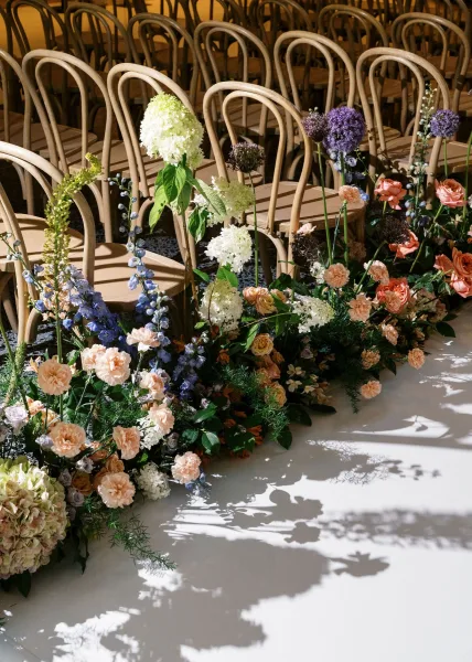 Ceremony aisle flowers in an aisle floral meadow along a white runner, with hydrangeas, roses, and bentwood chairs in sunlit indoor space