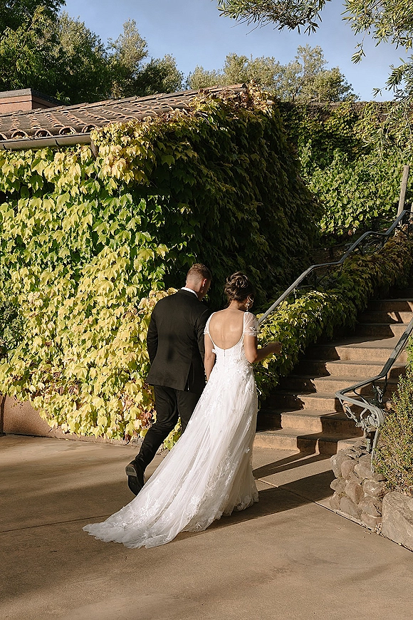 Couple portrait of bride and groom walking away up stone stairs, her open-back lace train flowing beside an ivy-covered wall