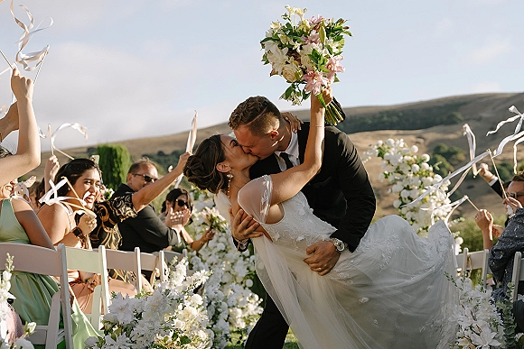 Wedding kiss as groom dips bride holding bouquet, veil flowing, guests waving ribbon wands along a white-flowered outdoor aisle with hills beyond