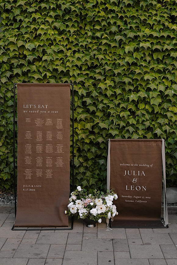 Wedding signage set with wedding welcome sign and seating chart on metal stands, framed by white and blush florals against an ivy wall backdrop