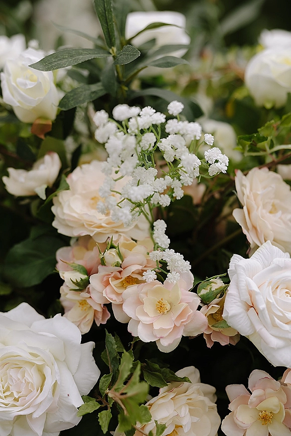 Wedding flowers featuring white roses and blush roses with baby’s breath accents and lush greenery, set against softly blurred florals and leaves