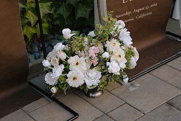 Wedding welcome sign with welcome sign floral arrangement of white roses, dahlias, blush blooms and greenery on metal stand on stone paver patio