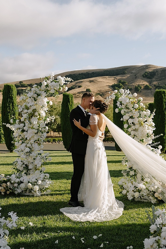 Wedding kiss portrait of bride and groom under a white floral arch, her veil flowing, with cypress trees and rolling hills behind