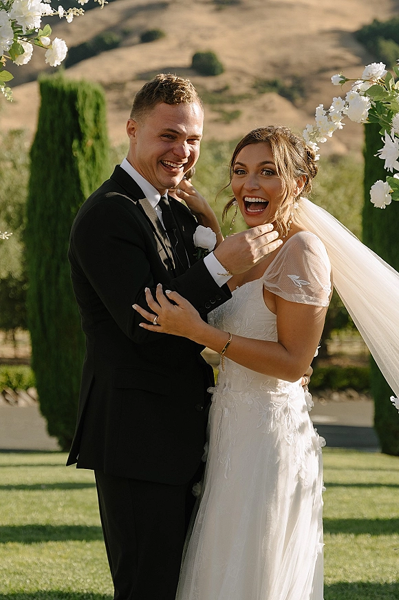Couple portrait of bride and groom laughing under a floral arch, her lace dress and veil flowing on a vineyard lawn with cypress trees
