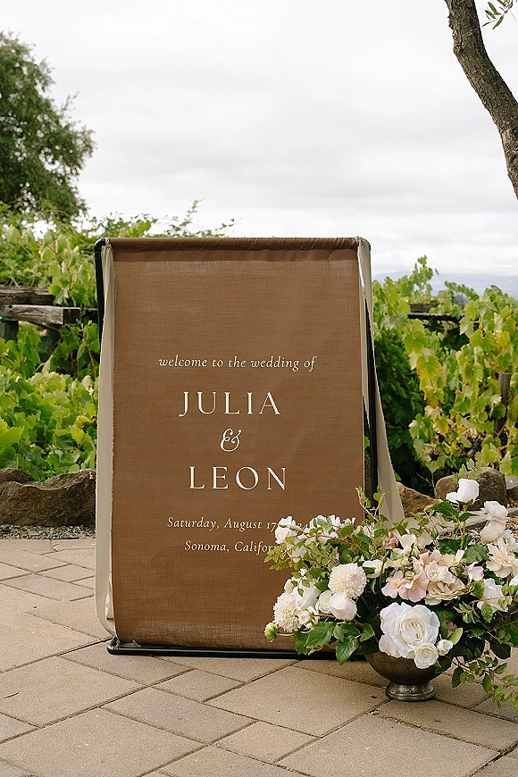 Wedding welcome sign on a fabric panel in a metal frame, tied with ribbon beside rose and greenery urn arrangement on a vineyard patio