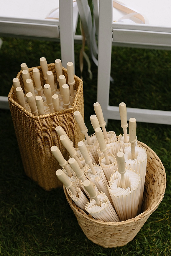 Wedding parasol basket filled with white paper parasols, wicker baskets and ribbon streamers on a grass lawn beside chair legs