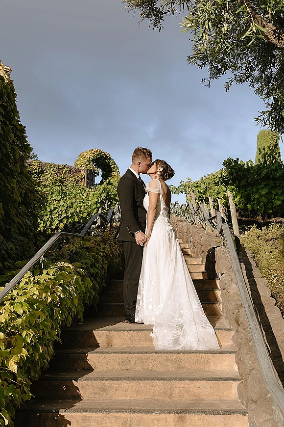 Wedding kiss portrait of bride and groom kissing on ivy-lined stone stairs, holding hands as her veil falls over lace dress beside wooden fence