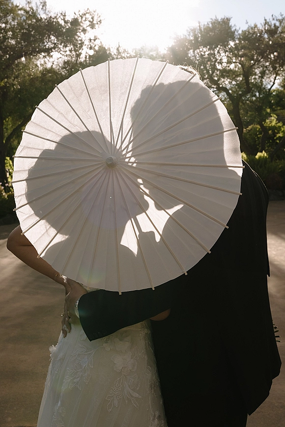 Wedding couple portrait of bride and groom kissing beneath a white paper parasol, backlit on a garden path with sun flare through trees