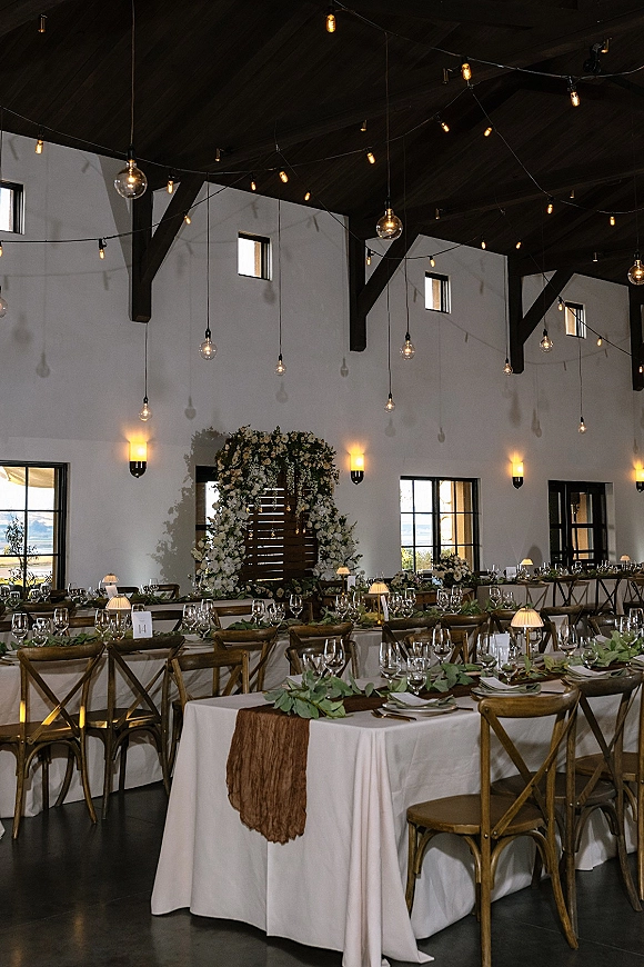 Reception tablescape with long banquet tables wedding styling, greenery garlands on brown runners, candlelit lamps, cross back chairs under string lights