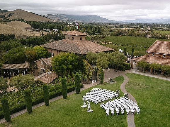 Outdoor ceremony setup with garden wedding ceremony seating in a curved semicircle, white folding chairs and floral aisle markers under string lights in a villa courtyard