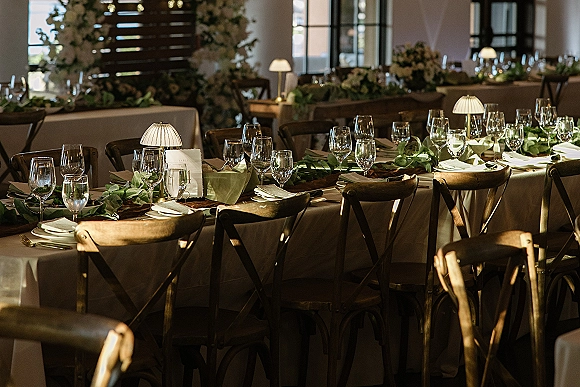 Reception tablescape with a greenery table runner, white linens, glassware, folded napkins, and small lamps on a long banquet table by windows