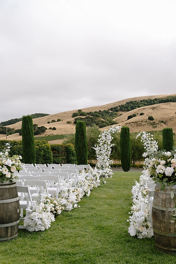 Ceremony aisle decor with outdoor wedding aisle flowers in white floral columns and barrel arrangements, on a lawn amid vineyard hills under overcast sky