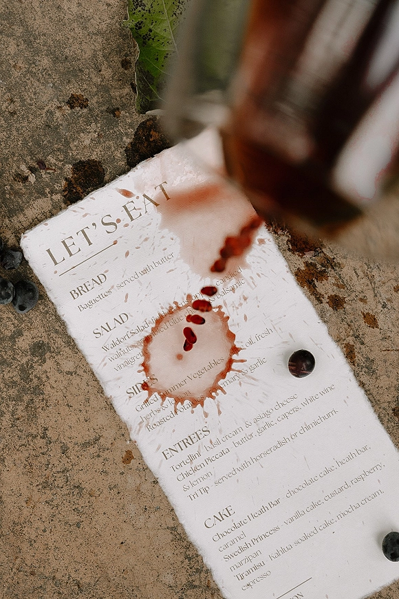 Wedding menu card with a reception menu card design, red wine spill and blueberries beside a wine glass on a stone surface with leaf