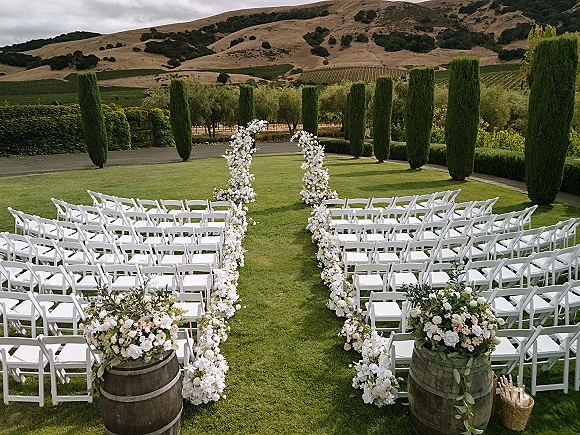 Ceremony setup with white folding chairs and aisle flowers leading to a white floral arch on a vineyard lawn with cypress trees
