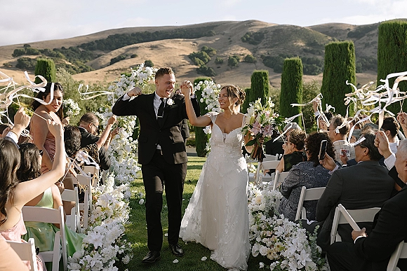 Wedding recessional as newlyweds walk down the aisle past cheering guests waving white ribbon wands, bride holding bouquet under a floral arch outdoors