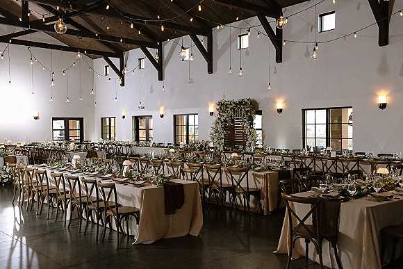 Reception tablescape with banquet table setup, linen cloths, cross back chairs, candles and small lamps beneath string lights in a bright hall