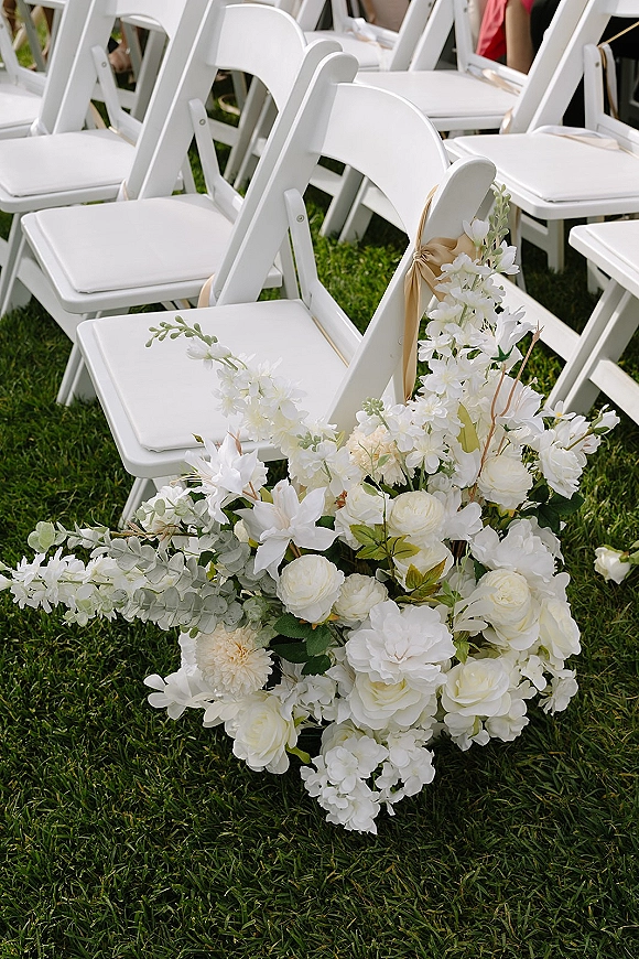 Ceremony aisle decor with outdoor ceremony chairs dressed in champagne sashes and a white-and-green floral arrangement on a grass lawn