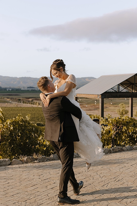 Wedding couple portrait of groom lifting bride in a lace dress and veil on a stone patio at a vineyard pavilion with string lights and mountains behind