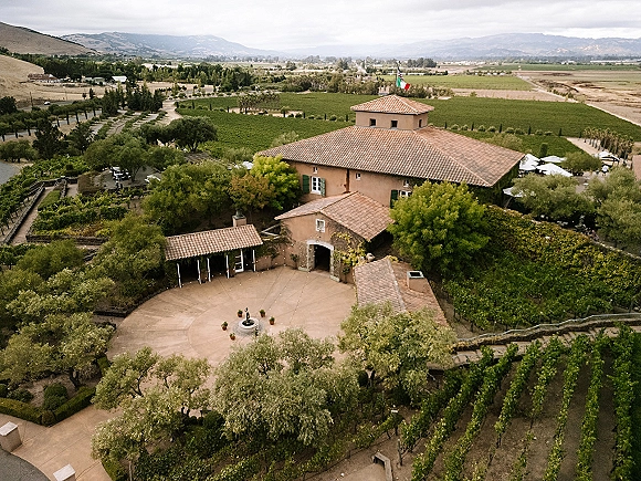 Vineyard wedding venue with a stone fountain in a courtyard, string lights and umbrellas, set against vineyard rows and mountains under clouds