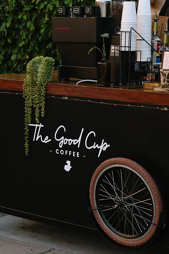 Wedding coffee cart with espresso machine and grinder, paper cups and syrup bottles, set against a green hedge wall on pavement