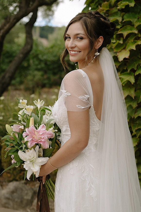 Bridal portrait of a bride looking over her shoulder, smiling in a lace gown and veil, holding a lily bouquet by an ivy wall