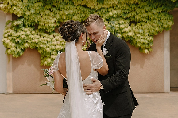 Wedding couple portrait of bride and groom embrace, her lace backless dress and veil against an ivy-covered stucco courtyard wall