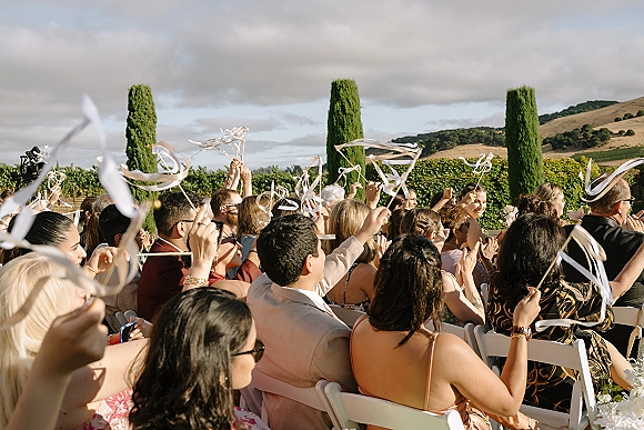 Wedding guests cheering with ribbon wands, white streamers on sticks raised over white folding chairs on an outdoor lawn with cypress trees