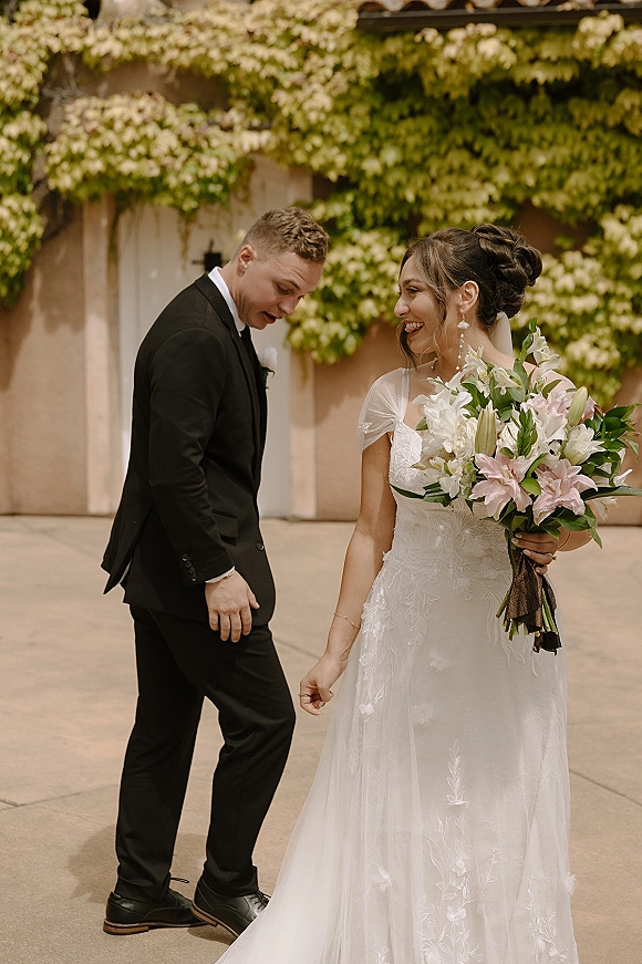 Couple portrait of bride holding bouquet laughing with groom in black tuxedo beside an ivy-covered stucco wall in a courtyard