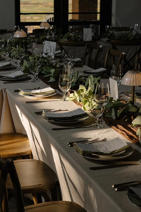 Reception tablescape with greenery garland table runner, white linens, menus, glasses and mini lamps on a long table by sunset-lit windows