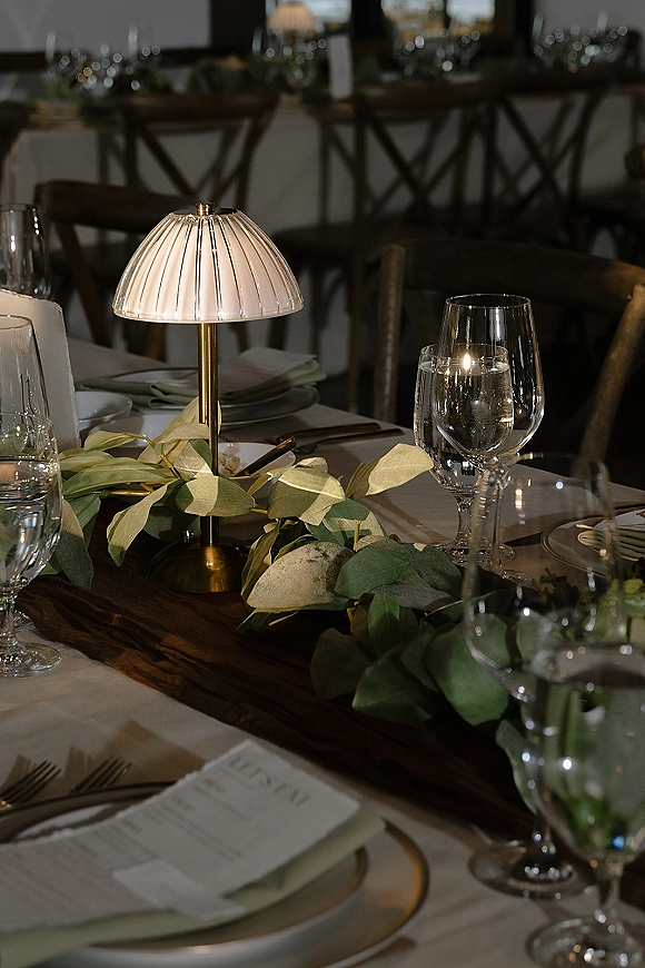 Reception tablescape with a wedding table lamp centerpiece on a greenery garland and wooden runner, set with menus, linens, and glassware at banquet tables