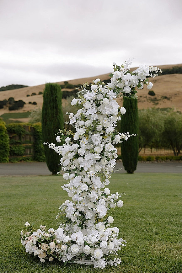 Wedding floral pillar with white wedding flower arrangement of roses, blossoms, and greenery on a stand against rolling hills and trees