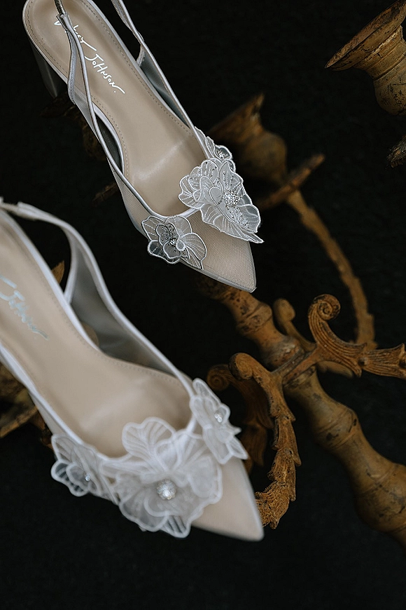 Wedding shoes with lace floral appliques and rhinestone accents, pointed-toe bridal slingback heels resting on an ornate gold chair