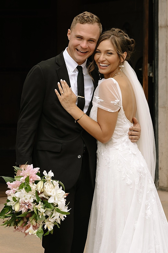 Couple portrait of bride and groom smiling, hugging by a dark doorway and stone wall, her veil and lace dress beside bouquet